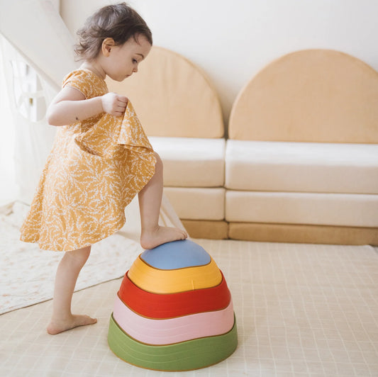 A young child in a yellow dress steps onto a set of Stacking Balance Stepping Stones – Vintage, arranged as a stack of four colorful nested stones to practice balance and coordination.