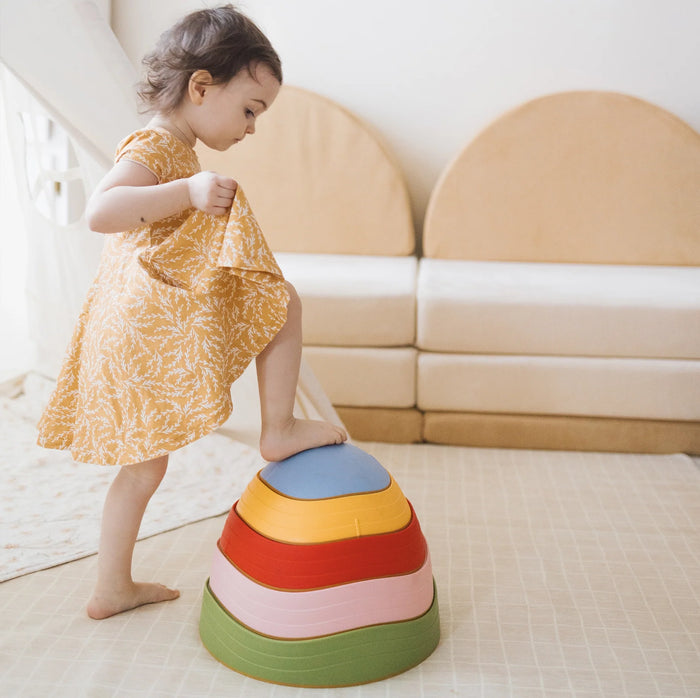 A young child in a yellow dress steps onto a set of Stacking Balance Stepping Stones – Vintage, arranged as a stack of four colorful nested stones to practice balance and coordination.