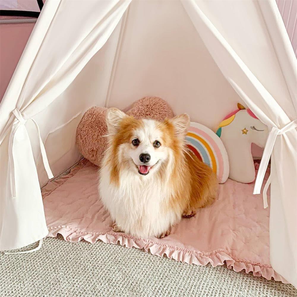 A fluffy dog sits inside a Cozy Kids Teepee Tent with Padded Mat on a pink ruffled mat with heart, rainbow, and unicorn-shaped pillows.