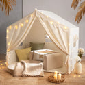 A beige Large Cozy Cabin Play Tent decorated with star lights, containing a small armchair, cushions, and a throw blanket on a woven floor mat to form a cozy reading nook.