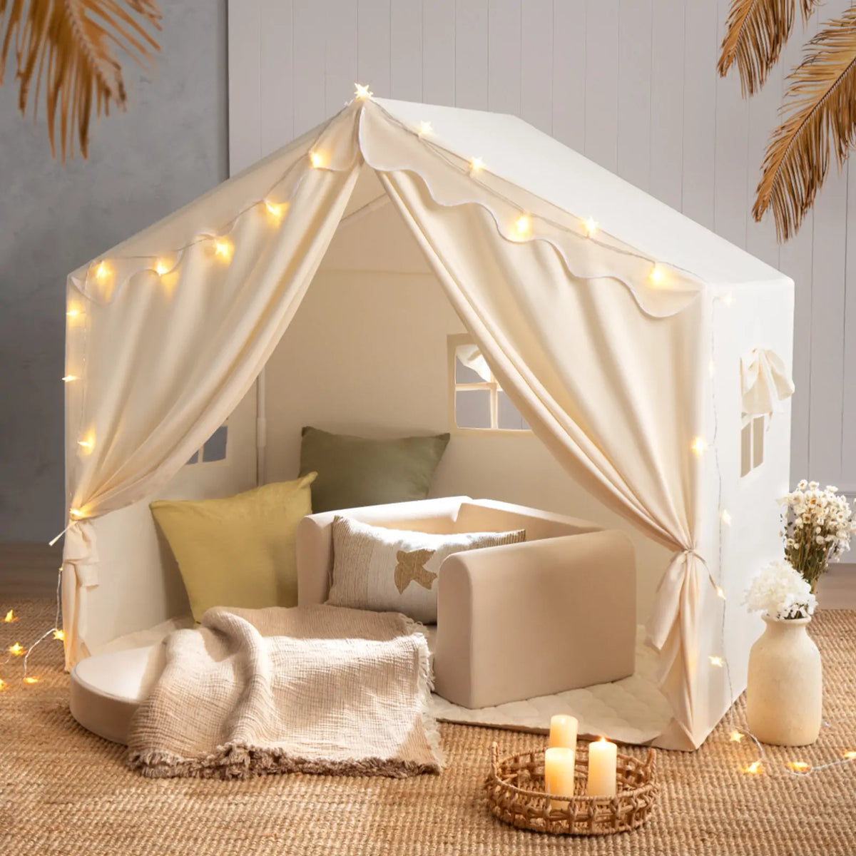 A beige Large Cozy Cabin Play Tent decorated with star lights, containing a small armchair, cushions, and a throw blanket on a woven floor mat to form a cozy reading nook.