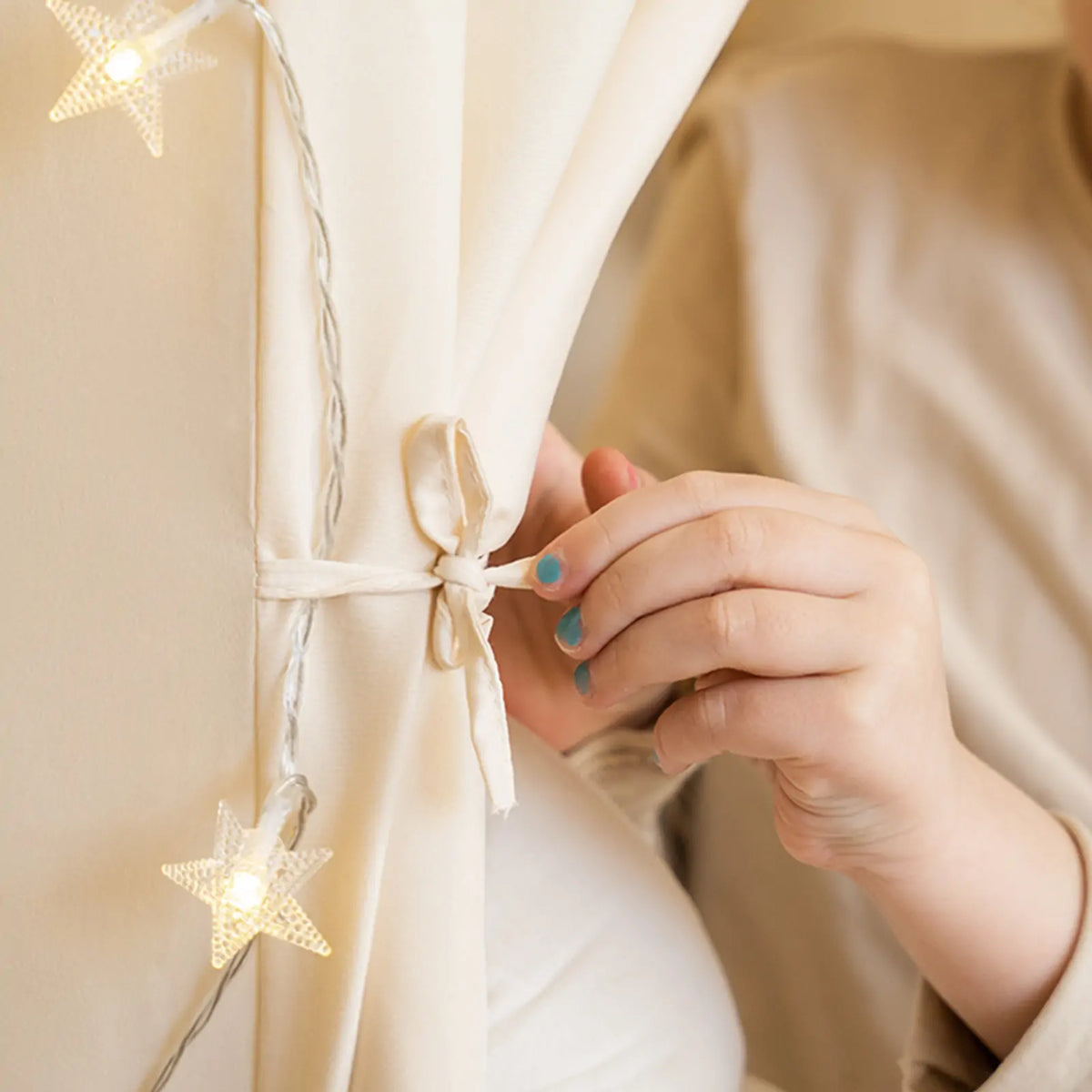 A hand ties back the beige fabric curtain of a Large Cozy Cabin Play Tent with Star Lights, which is accented with a string of illuminated star-shaped lights.