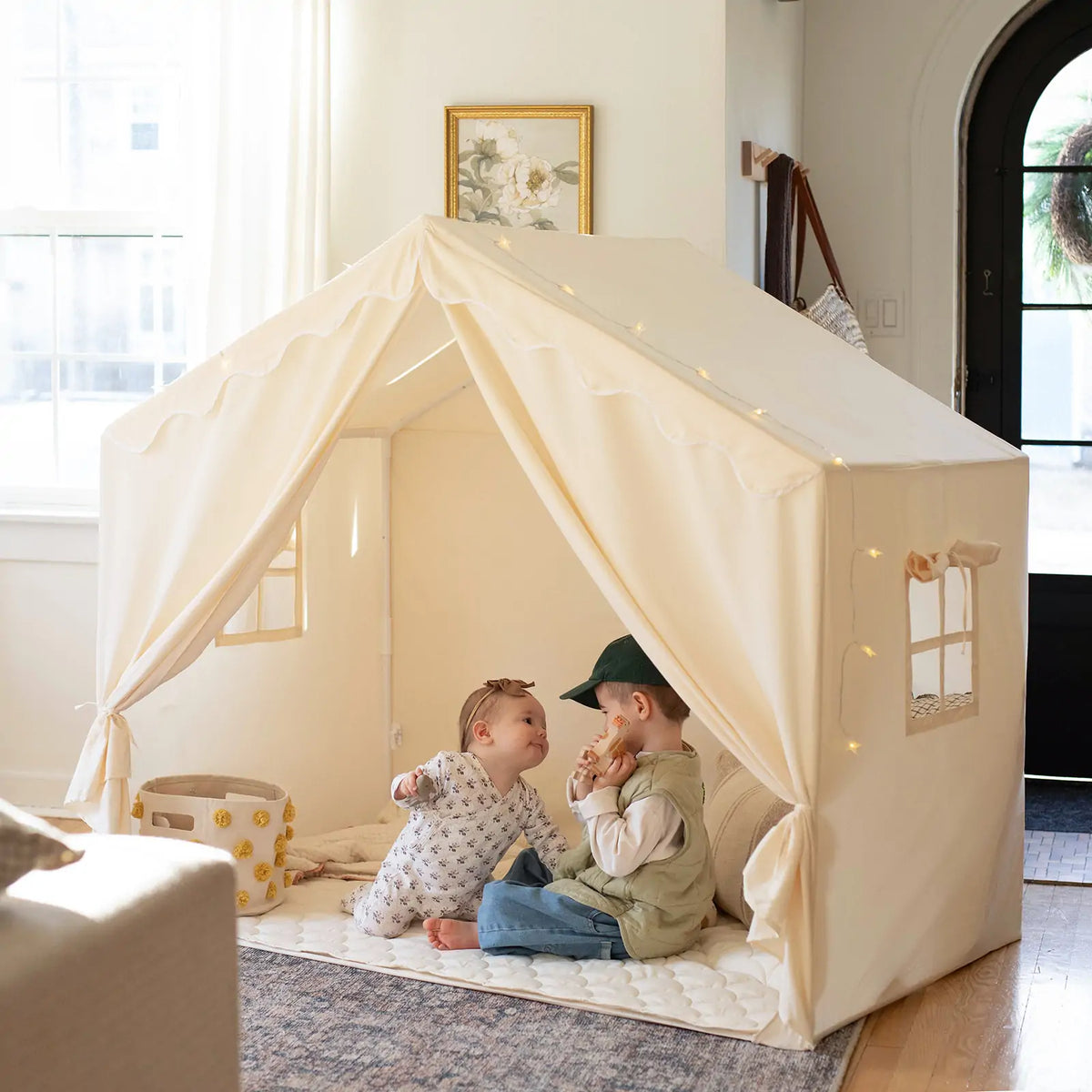 Two children sit inside a cream-colored Large Cozy Cabin Play Tent with Star Lights, which is decorated with string lights to create a cozy reading nook.
