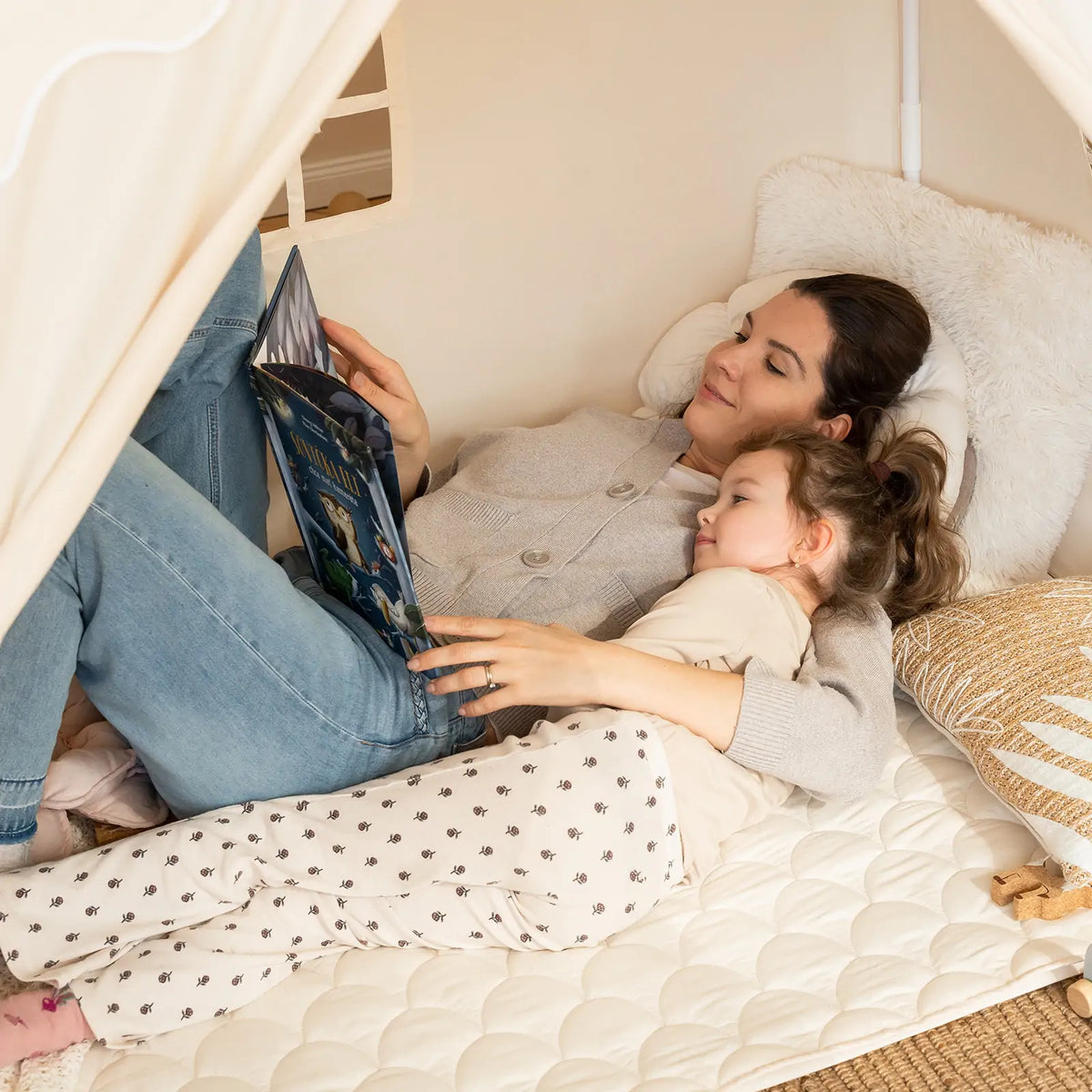 A mother and daughter lie together inside a Large Cozy Cabin Play Tent with Star Lights, sharing a picture book.