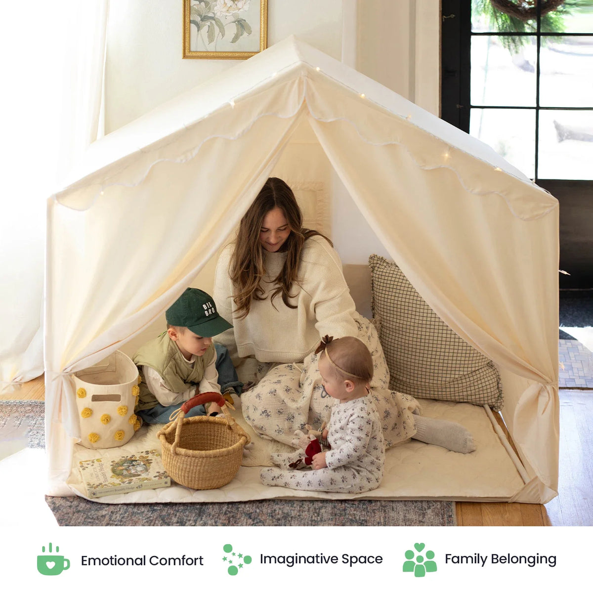A woman and two young children sit inside an off-white Large Cozy Cabin Play Tent with Star Lights, decorated with string lights to create a cozy reading nook.