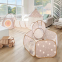A modern sunlit playroom features a beige and white star-themed Tiny Land® Discovery 5-in-1 Play Tunnel (no balls), a ball pit, and a cozy gray armchair. Wooden toys sit on light wood floors, with large windows showcasing lush greenery outside.