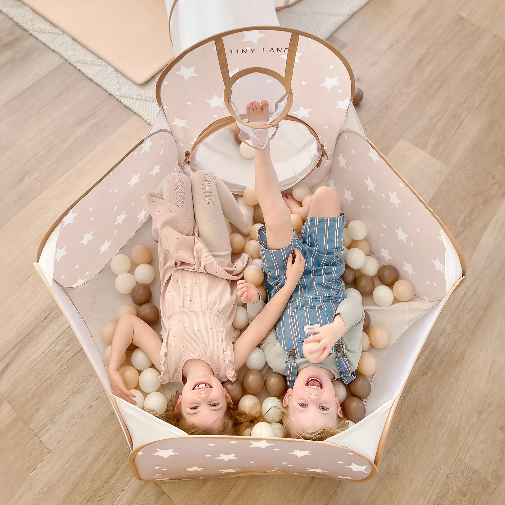 Two young children are smiling and laughing inside a beige, star-patterned playpen filled with soft, neutral-colored play balls, next to a Tiny Land® Discovery 5-in-1 Play Tunnel (no balls) on a wooden floor.