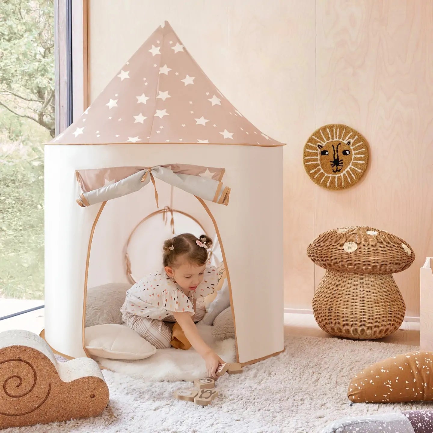 A young child plays inside a small, star-patterned play tent with the Tiny Land® Discovery 5-in-1 Play Tunnel With no Balls in a cozy room filled with plush toys, cushions, and wicker animal figures on a fluffy rug.