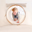A young child with blonde hair smiles while crawling through a soft, circular Tiny Land® Discovery 5-in-1 Play Tunnel With no Balls in a bright, white play area.