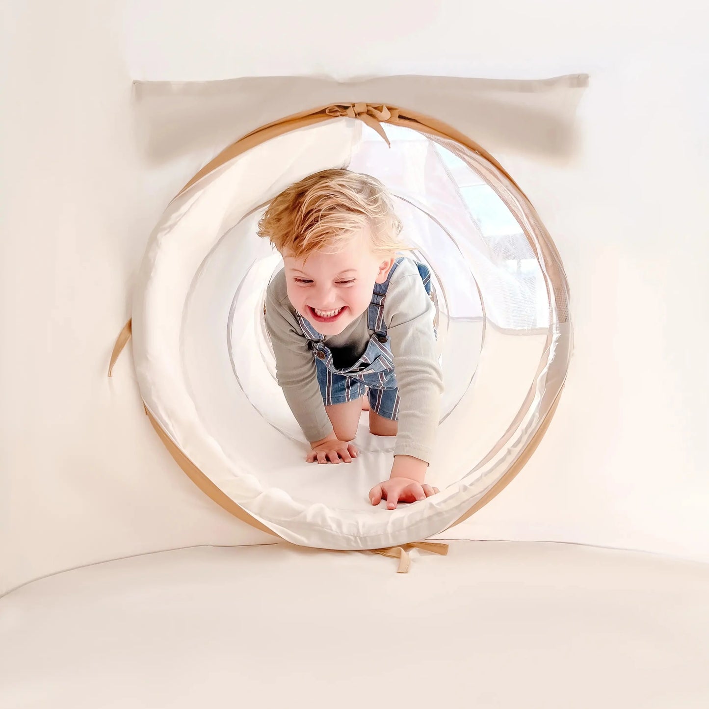 A young child with blonde hair smiles while crawling through a soft, circular Tiny Land® Discovery 5-in-1 Play Tunnel With no Balls in a bright, white play area.