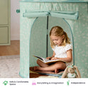 A young girl sits inside a sage green floral Starry Indoor Play Tent for Toddlers reading a book, with a stuffed rabbit sitting beside her.