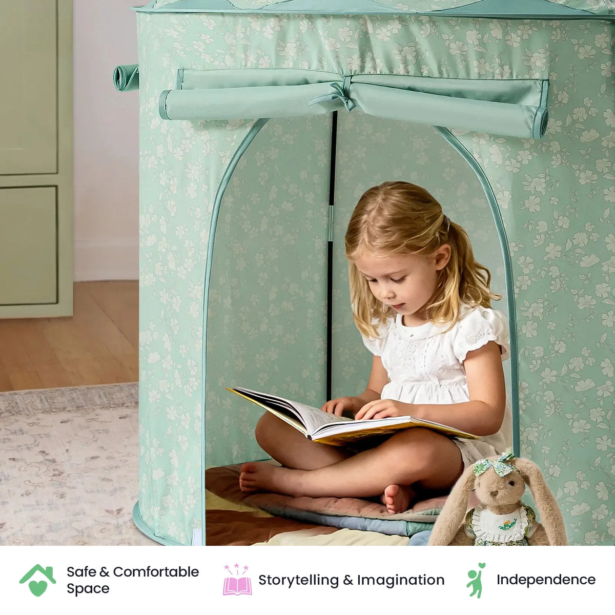 A young girl sits inside a sage green floral Starry Indoor Play Tent for Toddlers reading a book, with a stuffed rabbit sitting beside her.