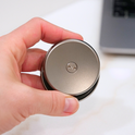 A hand holds the ONO Slider Titanium, a small, round fidget toy with ONO engraved on top; a laptop appears blurred in the background.