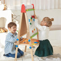 Two young children enjoy the Tiny Land® 3-in-1 Art Easel for Kids, attaching magnetic letters on its whiteboard. The boy sits and smiles as the girl stands to reach up. The bright, cozy room features light-colored decor.