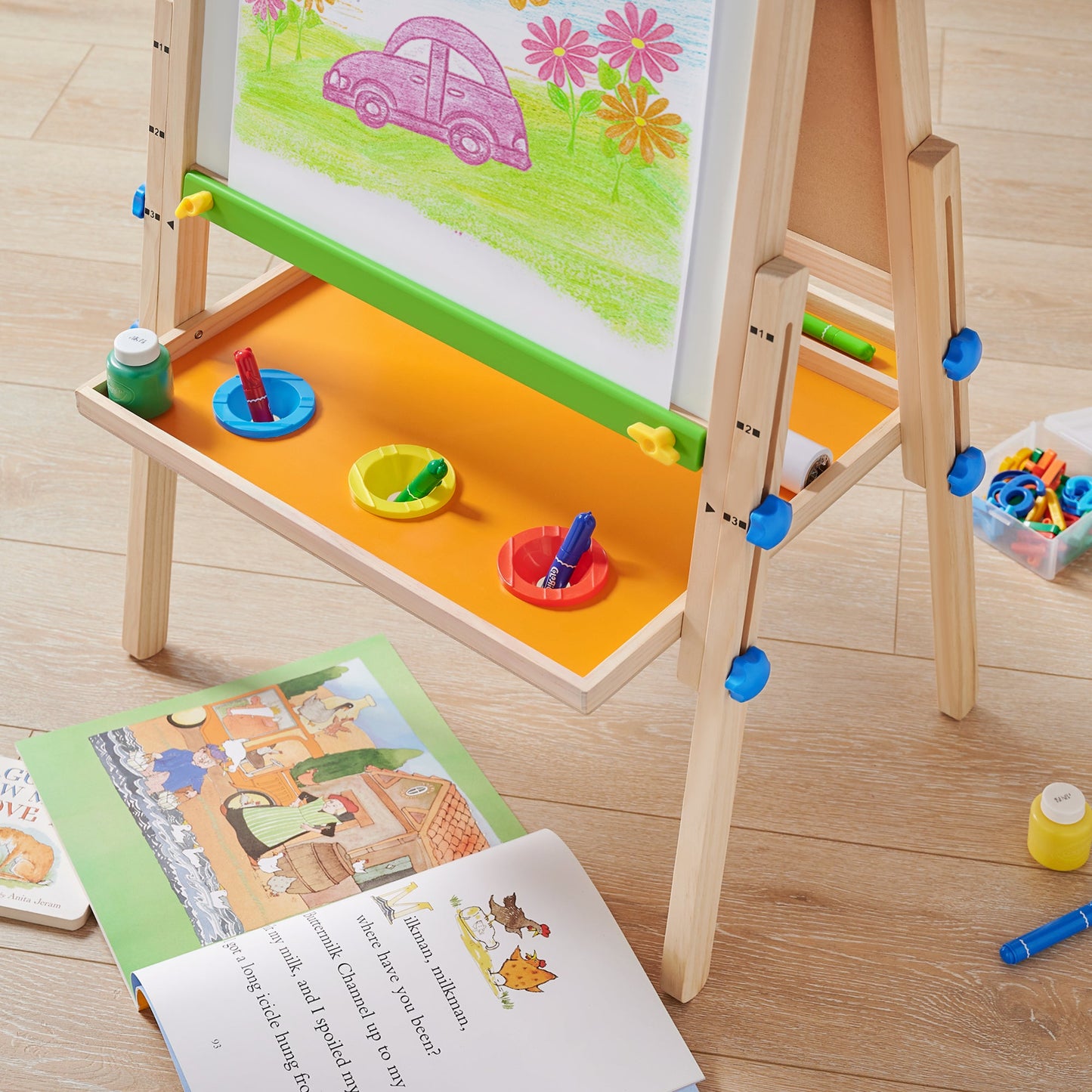 The Tiny Land® 3-in-1 Art Easel for Kids displays a purple car and bright flowers. Markers and paint rest on its shelf, while open picture books and toys lie scattered on the light wood floor below.