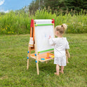 A young child stands on grass, drawing with crayons on the Tiny Land® 3-in-1 Art Easel for Kids while holding a stuffed toy. The child faces away from the camera, surrounded by tall grass and green trees outdoors.