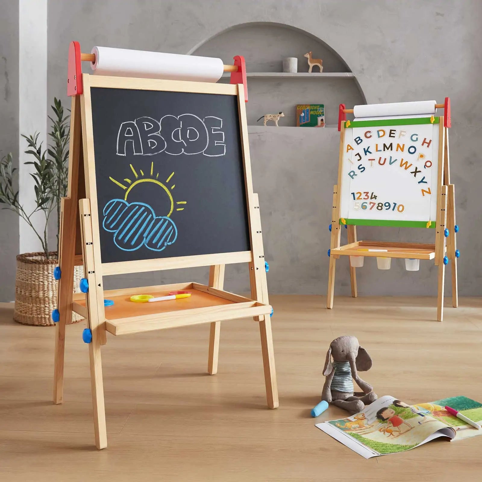 The Tiny Land® 3-in-1 Art Easel for Kids, a double-sided wooden easel with a chalkboard showing colorful drawings and ABCDE letters, stands in a playroom with art supplies and a plush bunny nearby. Another easel is visible in the background.