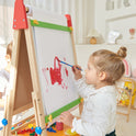 A young girl with light brown hair paints on the Tiny Land® 3-in-1 Art Easel for Kids in a bright, cozy room. Art supplies are scattered nearby, and another child is partially visible beside her.