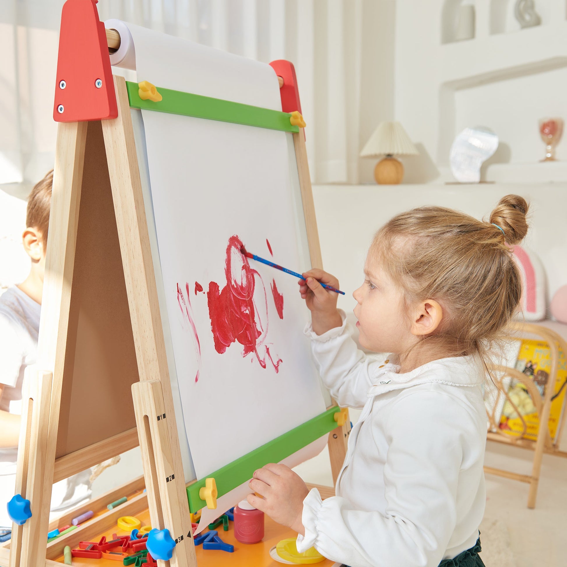 A young girl with light brown hair paints on the Tiny Land® 3-in-1 Art Easel for Kids in a bright, cozy room. Art supplies are scattered nearby, and another child is partially visible beside her.