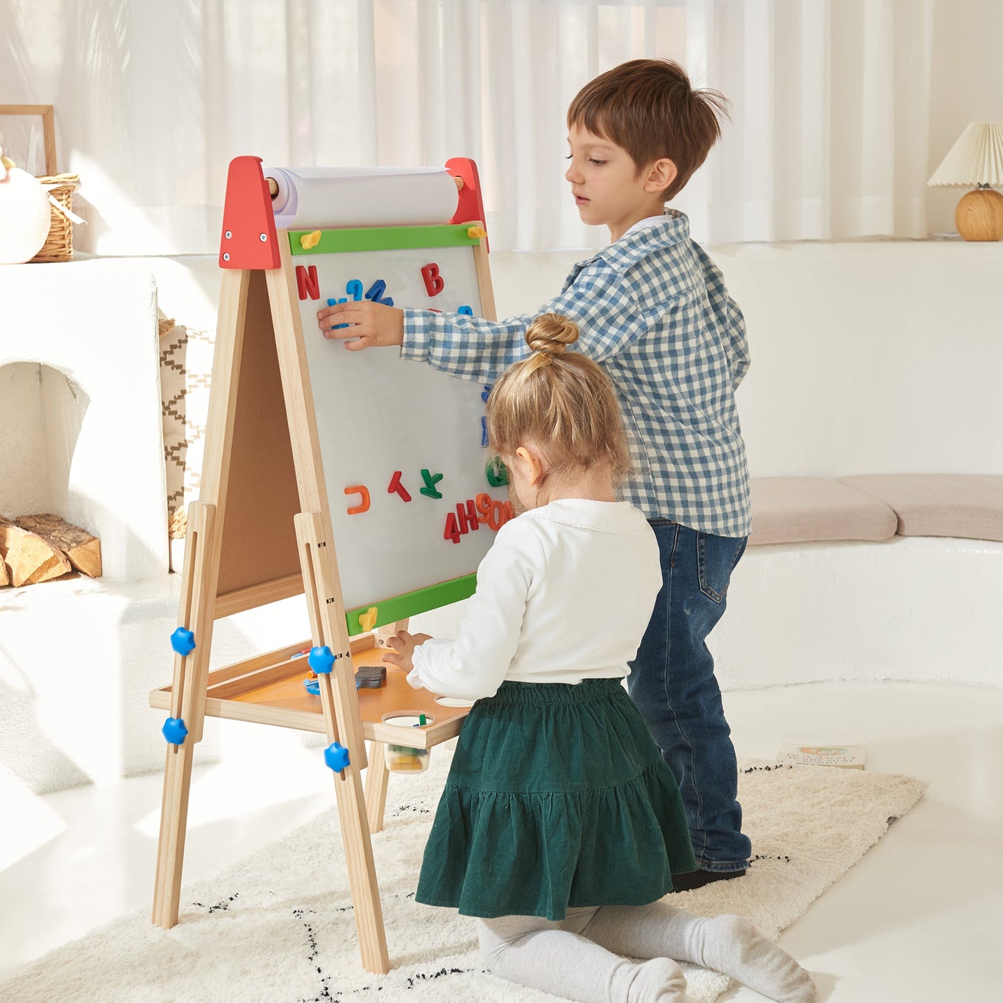 A boy and girl play with colorful magnetic letters on the Tiny Land® 3-in-1 Art Easel for Kids in a bright, cozy room. The boy stands while the girl kneels on the rug, both focused on arranging the magnets.