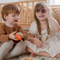 Two children sit on a rug playing with a Wooden Play Food Set & Storage Rack Bundle, one holding a carrot while the other looks through a toy donut held over their eye.