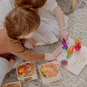 Two children sit on a rug playing with a Wooden Play Food Set & Storage Rack Bundle on a Farmers Market mat, with additional pieces organized in a storage rack.