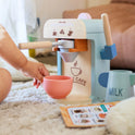A toddler reaches for a pink play cup under a Wooden Play Coffee Maker Set, with a toy milk pitcher and menu card from the barista playset nearby.