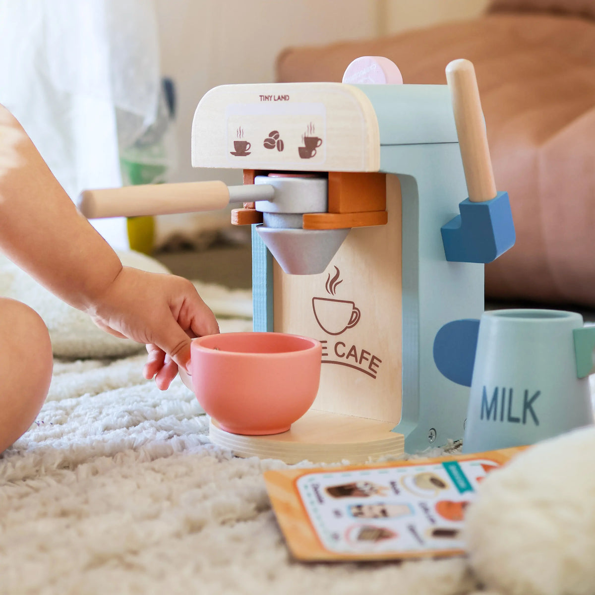 A toddler reaches for a pink play cup under a Wooden Play Coffee Maker Set, with a toy milk pitcher and menu card from the barista playset nearby.