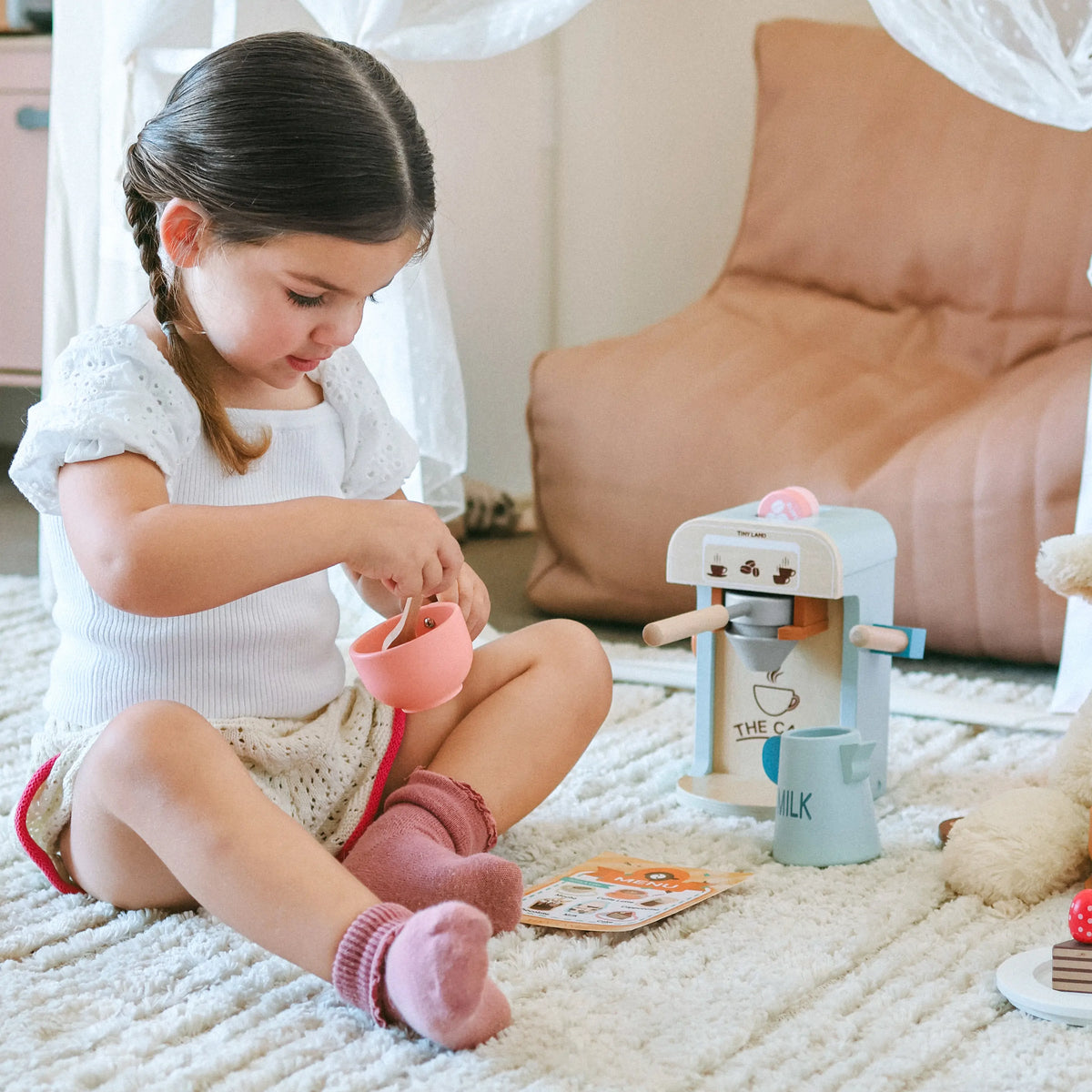A child sitting on a rug plays with a Wooden Play Coffee Maker Set, holding a pink cup as part of the toy.