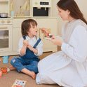 A woman feeds a piece of toy cake to a child sitting on the floor in front of a Wooden Play Coffee Maker Set.