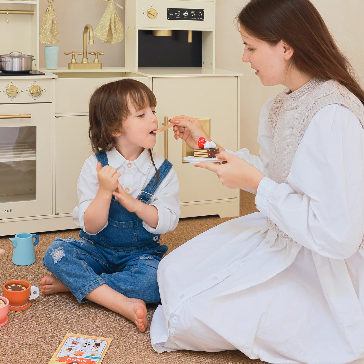 A woman feeds a piece of toy cake to a child sitting on the floor in front of a Wooden Play Coffee Maker Set.