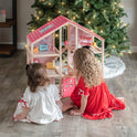 Two young children in festive pajamas play imaginative games with the Tiny Land® Sweetwood Love Dollhouse without Dolls on a wooden floor, set in front of a Christmas tree glowing with string lights.