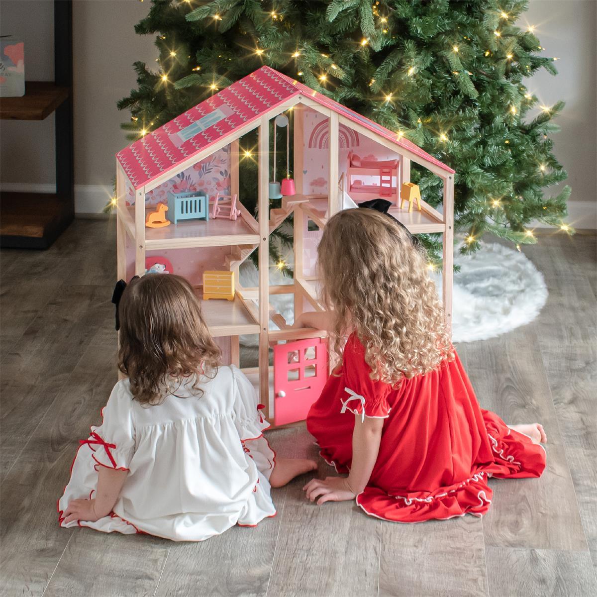 Two young children in festive pajamas play imaginative games with the Tiny Land® Sweetwood Love Dollhouse without Dolls on a wooden floor, set in front of a Christmas tree glowing with string lights.