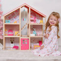 A young girl in a pink dress smiles beside the Tiny Land® Sweetwood Love Dollhouse without Dolls, a classic toy with miniature furniture and pink floral wallpaper that inspires hours of imaginative play.