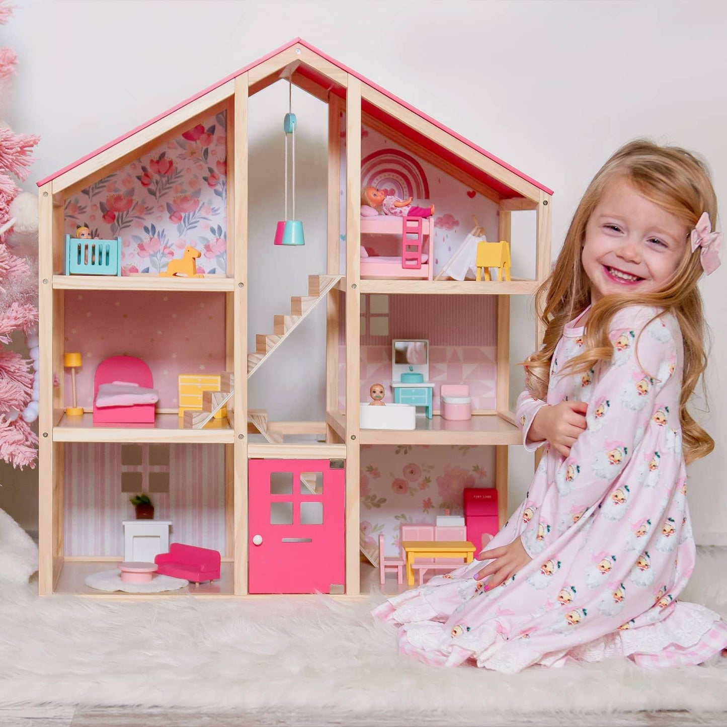 A young girl in a pink dress smiles beside the Tiny Land® Sweetwood Love Dollhouse without Dolls, a classic toy with miniature furniture and pink floral wallpaper that inspires hours of imaginative play.