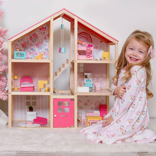 A young girl in a pink dress smiles beside the Tiny Land® Sweetwood Love Dollhouse without Dolls, a classic toy with miniature furniture and pink floral wallpaper that inspires hours of imaginative play.