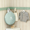 A round, sage green Starry Indoor Play Tent for Toddlers carrying case hangs on a wall hook next to a beige tote bag and a grey knit cardigan, with a small ceramic deer figure on the surface below.