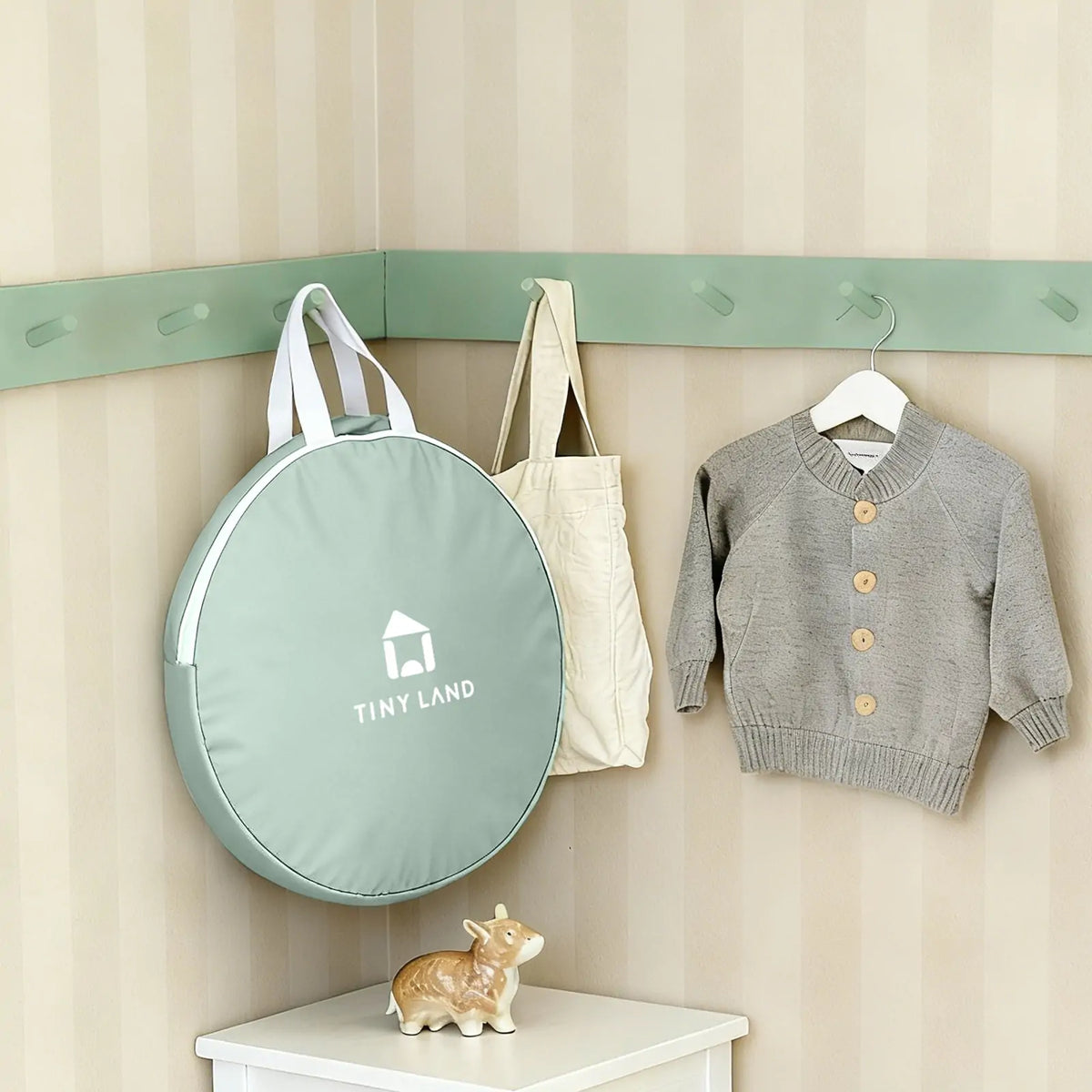 A round, sage green Starry Indoor Play Tent for Toddlers carrying case hangs on a wall hook next to a beige tote bag and a grey knit cardigan, with a small ceramic deer figure on the surface below.