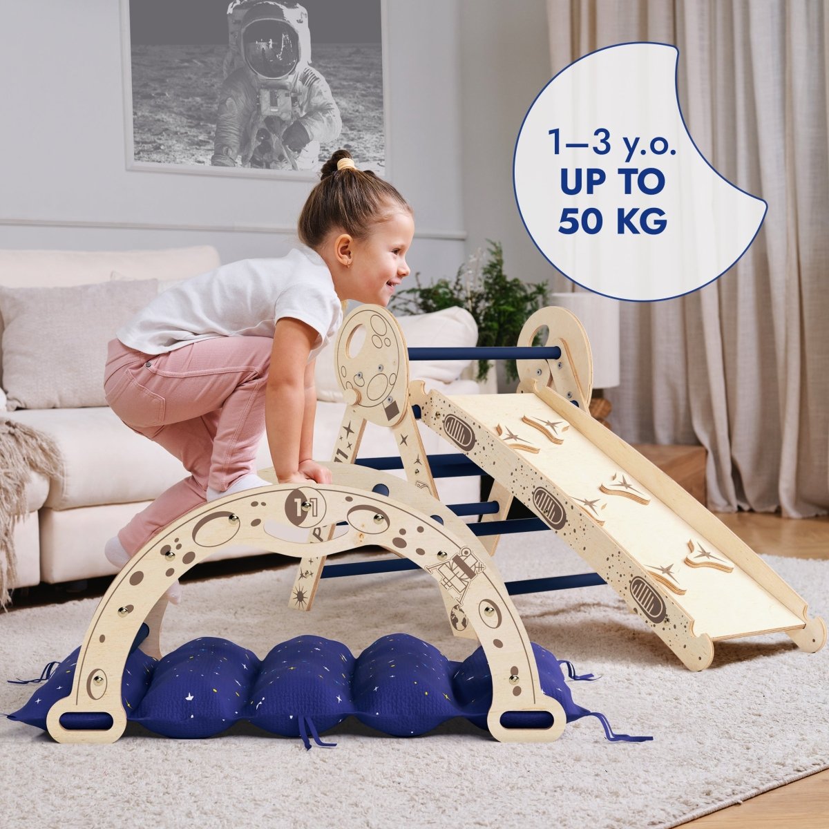 A young child climbs on the Apollo 4in1 Climbing Set: Pikler Triangle, Arch, Slide Board, and Cushion for Little Toddlers Astronauts (1–3 Years), a wooden indoor Montessori climbing set in a living room. Text reads 1–3 y.o. UP TO 50 KG. A picture of an astronaut decorates the wall in the background, adding to the fun atmosphere for toddler climbing toys.