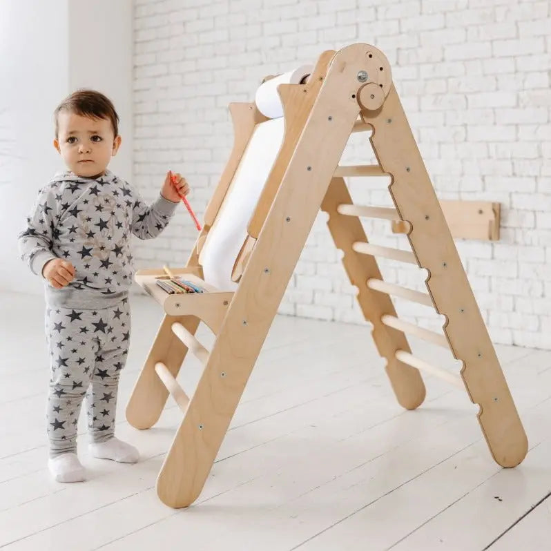 A toddler in star-patterned pajamas stands by a Goodevas Art Addition to the Triangle Ladder, which features a roll of paper and crayons, in a bright, white room.