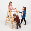 Three young children draw and color together on a roll of paper attached to a wooden easel. One child stands confidently on the Triangle Ladder, while the others stand and draw below. This adjustable art station, the Art Addition to the Triangle Ladder, provides a creative environment against a plain white background.