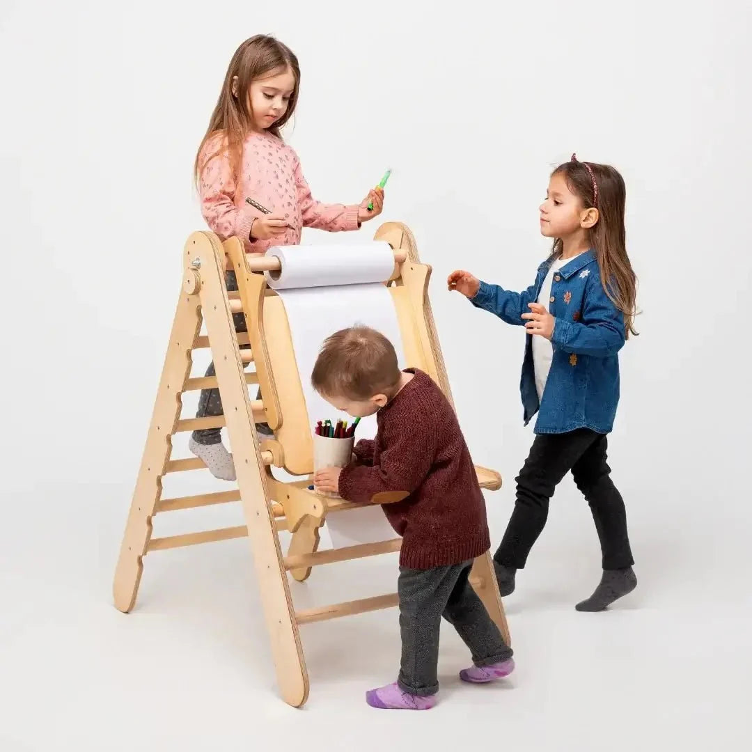 Three young children draw and color together on a roll of paper attached to a wooden easel. One child stands confidently on the Triangle Ladder, while the others stand and draw below. This adjustable art station, the Art Addition to the Triangle Ladder, provides a creative environment against a plain white background.