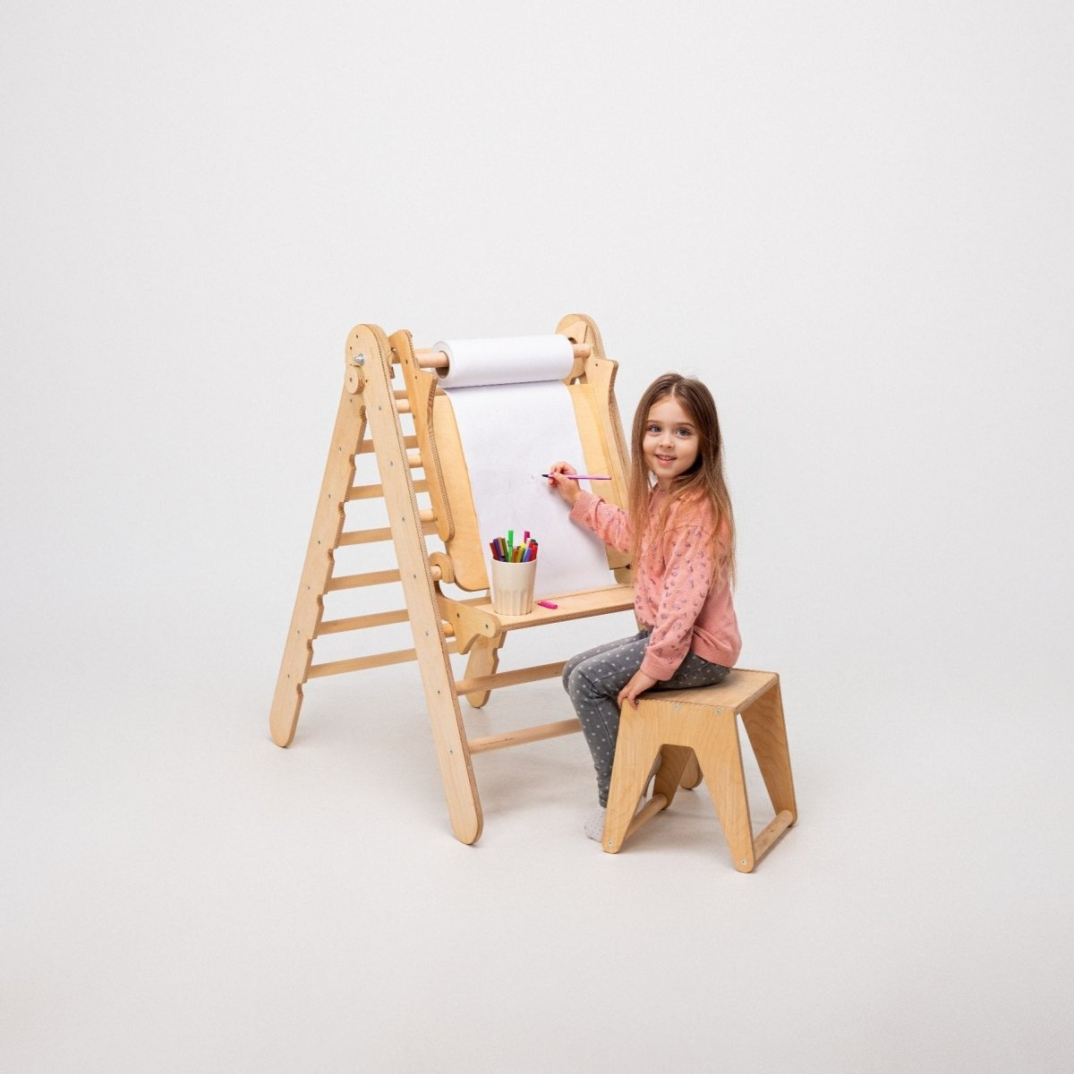 A young girl sits on a wooden stool, drawing on a large, vertical paper roll attached to the Art Addition to the Triangle Ladder by Goodevas. Colored markers are in a cup on the easel, while the triangle ladder stands nearby. The background is plain white.