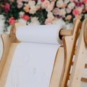 The Art Addition to the Triangle Ladder, an adjustable wooden easel, holds a roll of white paper adorned with faint colorful scribbles, set against a softly blurred background of pink and white flowers.
