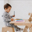 A toddler in star-patterned pajamas sits at a small wooden desk that doubles as an adjustable art station, holding a counting stick. Several colorful sticks and a notebook rest on the desk in front of them, hinting at creative possibilities inspired by the Art Addition to the Triangle Ladder.