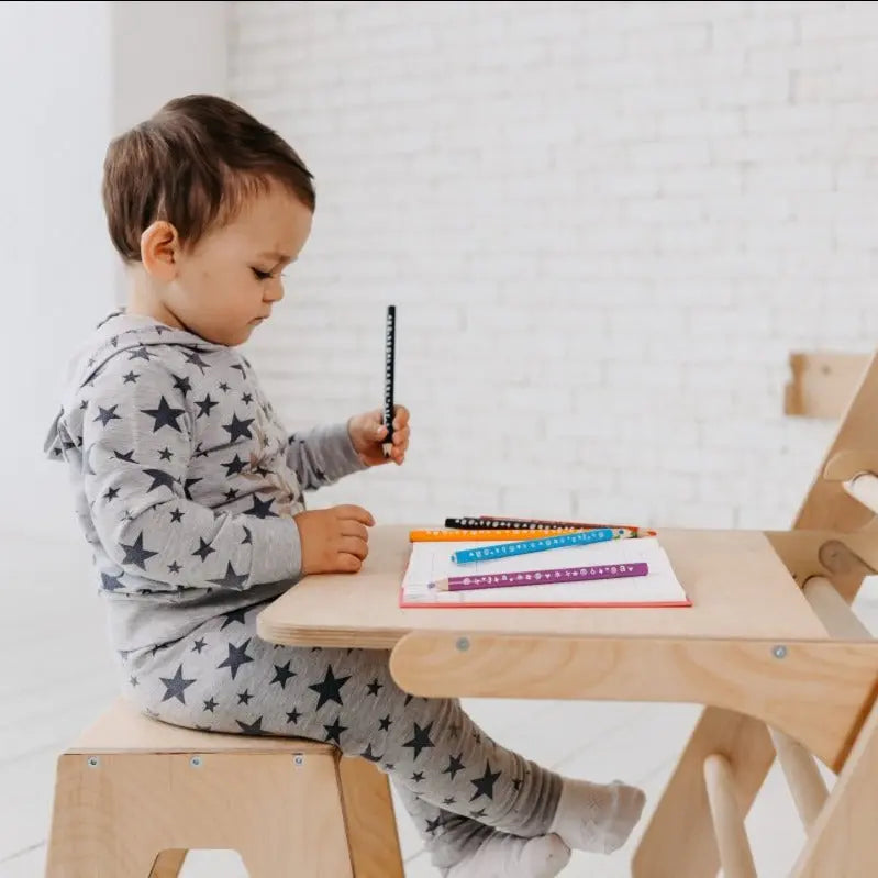 A toddler in star-patterned pajamas sits at a small wooden desk that doubles as an adjustable art station, holding a counting stick. Several colorful sticks and a notebook rest on the desk in front of them, hinting at creative possibilities inspired by the Art Addition to the Triangle Ladder.