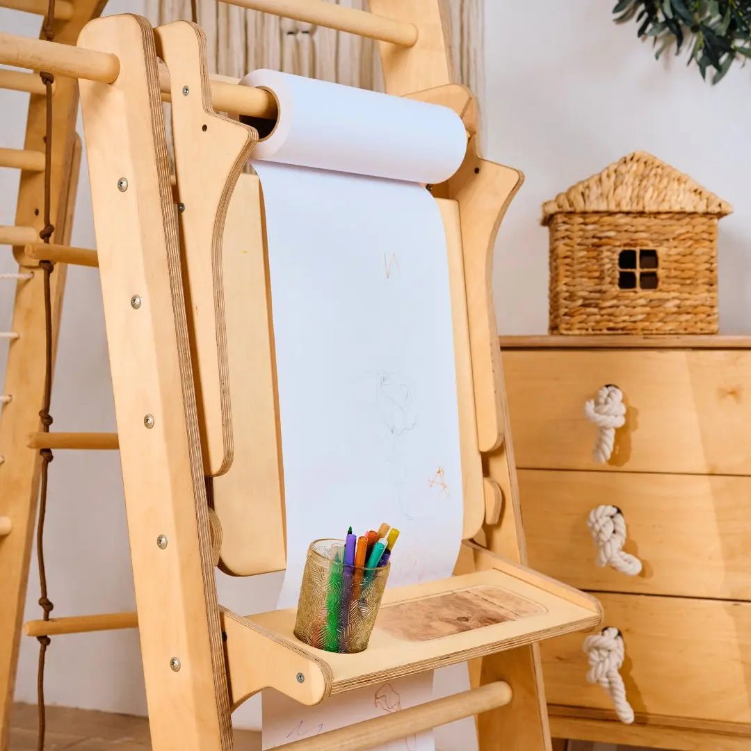 A wooden easel with a roll of white paper, some faint drawings, and a small tray holding colored pencils creates an inviting Art Addition to the Triangle Ladder by Goodevas in the space. An adjustable art station sits nearby, while a wicker house and wooden drawers in the background contribute to a cozy art environment.
