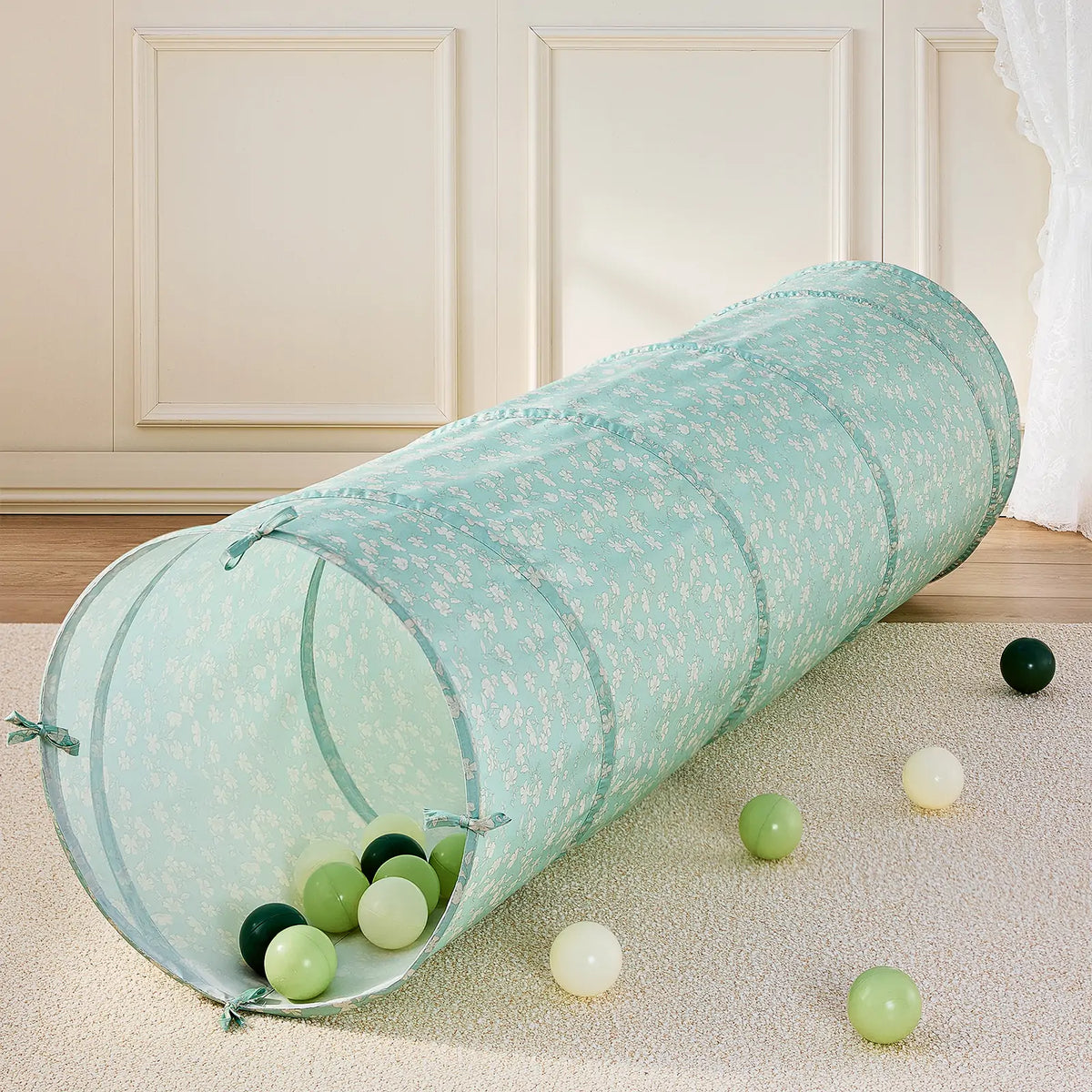 A long, collapsible green Adventure Crawl Tunnel (68″) patterned with small white flowers, resting on a carpet with several scattered play balls inside and around it.