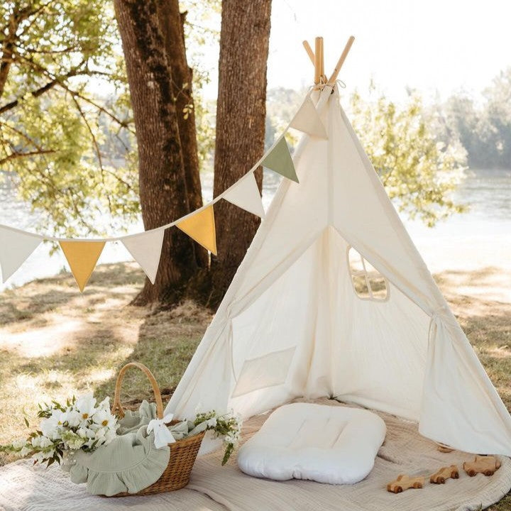 A white Cozy Kids Teepee Tent with Padded Mat featuring bunting flags and a picnic basket of flowers, set up outdoors near a river.