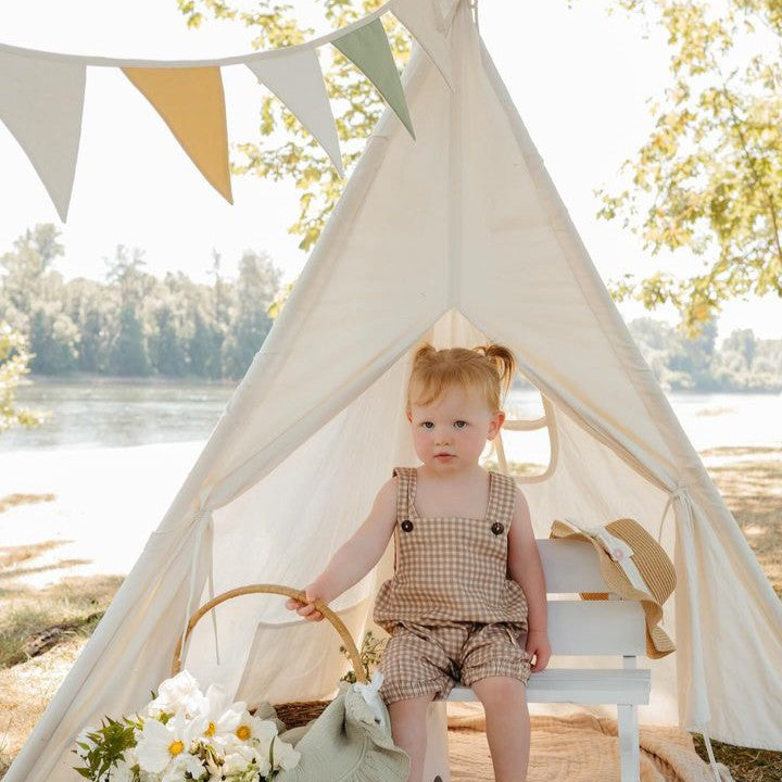 A toddler sits inside a Cozy Kids Teepee Tent with Padded Mat decorated with pennant flags, with a basket of white flowers at their side on a padded floor mat.